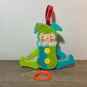 May include: Vintage wooden pull-along toy featuring a seated clown with a green hat and turquoise and green body. The clown has a red ribbon for hanging and an orange pull ring. The toy is designed with a smiling face and yellow accents.