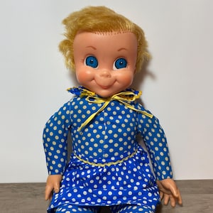 May include: A vintage doll with blonde hair and blue eyes. The doll is wearing a blue outfit with white polka dots, a yellow collar, and a yellow bow. The doll's skin is a light peach color.