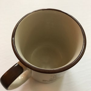 K&ouml;nnte beinhalten: Eine braune Keramiktasse mit wei&szlig;em Innenraum und braunem Henkel. Die Tasse hat einen braunen Rand und ein braunes Band um den Boden. Die Tasse ist leer.