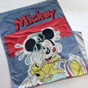 Pode incluir: Uma toalha de praia azul e vermelha com uma imagem de desenho animado do Mickey Mouse usando &oacute;culos de sol. A toalha tem a palavra "Mickey" impressa em letras vermelhas.