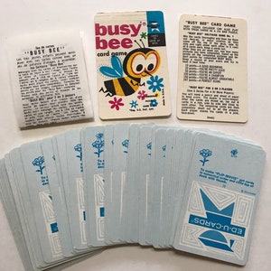 以下が含まれることがあります： 箱に青と黄色の蜂のイラストが描かれた、ビンテージのカードゲーム「ビジービー」。ゲームには、青と白の花のイラストと青と白の幾何学模様が描かれたカードデッキが含まれています。