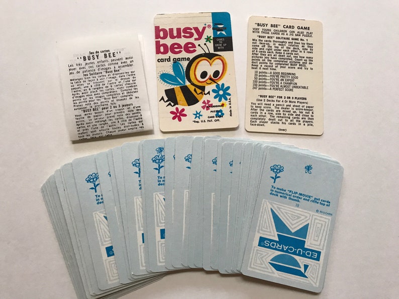 以下が含まれることがあります： 箱に青と黄色の蜂のイラストが描かれた、ビンテージのカードゲーム「ビジービー」。ゲームには、青と白の花のイラストと青と白の幾何学模様が描かれたカードデッキが含まれています。