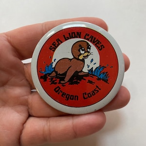 May include: A white button with a red and white design. The button features a cartoon sea lion with the text "Sea Lion Caves Oregon Coast".