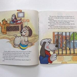 May include: A book illustration of a group of grey dogs in a pound. The dogs are looking out of a cage at a boy in a wheelchair. The text in the image reads, "Hey, everybody. Cooler whispered. "I think we've got a live one out there! You all know what to do."