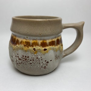 May include: A handmade ceramic mug with a unique speckled design. The mug features a light brown rim and handle, with a textured, speckled gray body. A band of brown and yellow glaze adds a decorative touch. The mug is designed for hot beverages.