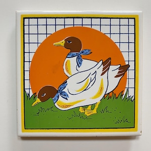 May include: A white ceramic tile with a yellow border and a grid pattern. The tile features a colourful illustration of two ducks with blue bandanas in front of an orange sun and green grass.
