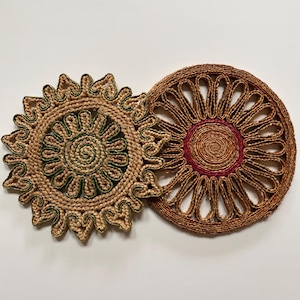 May include: Two circular woven placemats. One is light brown with green detailing, the other is a darker brown with a red centre. The placemats feature a sunburst design and are made from natural fibres.