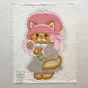 以下が含まれることがあります： ピンクのドット柄の帽子とドレスを着て花を持っている漫画の猫が描かれた布パネル。猫はオレンジ色の毛皮と緑色の目をしています。パネルには指示と「LEAVE OPEN FOR STUFFING」というテキストが含まれています。