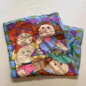 May include: A Cabbage Patch Kids quilt featuring a group of the dolls. The blanket has a blue border with green floral accents. The dolls have various hair colours and wear different outfits. The words "Cabbage Patch Kids" are visible on a hat.