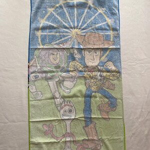 Puede incluir: Una toalla de playa verde y azul con una imagen de dibujos animados de Woody, Buzz Lightyear y Forky de la franquicia de pel&iacute;culas Toy Story. La toalla presenta una noria en el fondo.