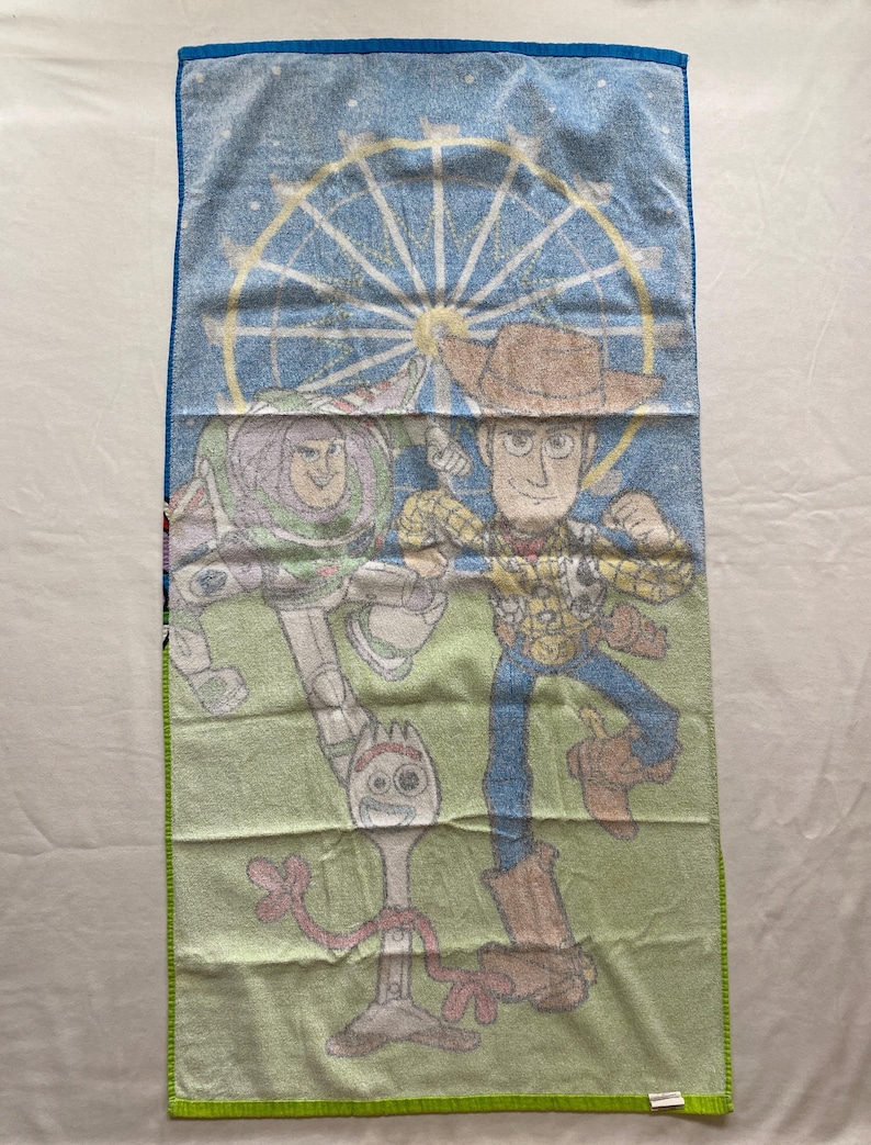 Puede incluir: Una toalla de playa verde y azul con una imagen de dibujos animados de Woody, Buzz Lightyear y Forky de la franquicia de pel&iacute;culas Toy Story. La toalla presenta una noria en el fondo.
