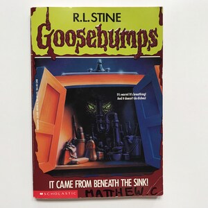 Op de afbeelding: Een Goosebumps-boekomslag met een rode titel met druppelend bloed, "Goosebumps", en een gele achtergrond. De omslagillustratie toont een gootsteen met de groene ogen van een monster die onder de gootsteen vandaan kijken. De titel van het boek is "It Came From Beneath The Sink!" met de naam van de auteur, "R.L. Stine", bovenaan. Het boek is uitgegeven door Scholastic.