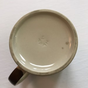 K&ouml;nnte beinhalten: Die Unterseite einer wei&szlig;en Keramiktasse mit braunem Henkel. Die Tasse hat eine kleine Markierung auf der Unterseite, die "AG PAK" lautet.