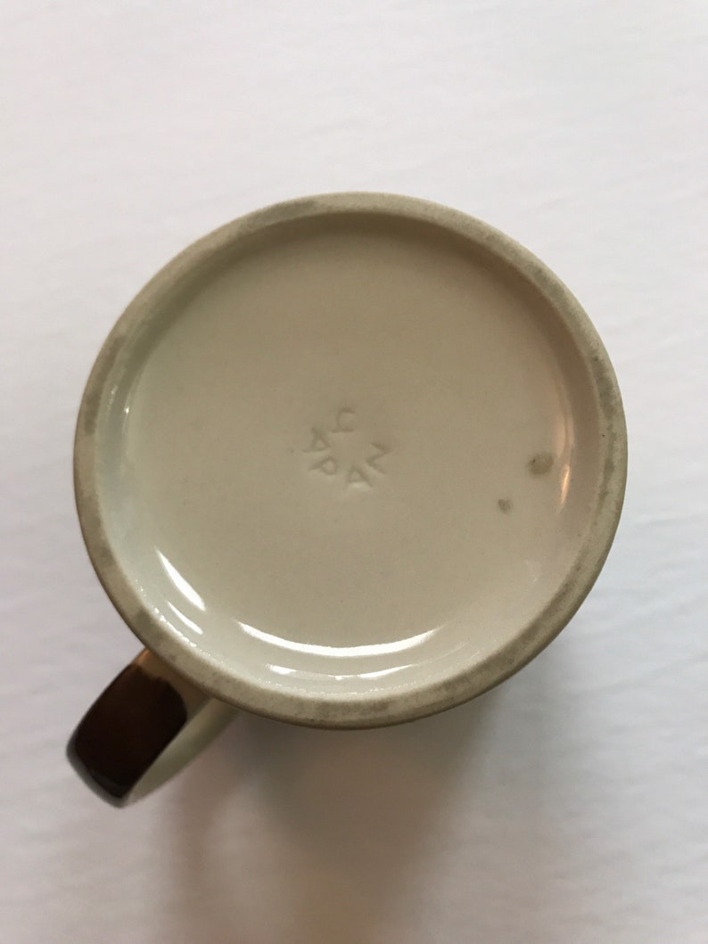 K&ouml;nnte beinhalten: Die Unterseite einer wei&szlig;en Keramiktasse mit braunem Henkel. Die Tasse hat eine kleine Markierung auf der Unterseite, die "AG PAK" lautet.