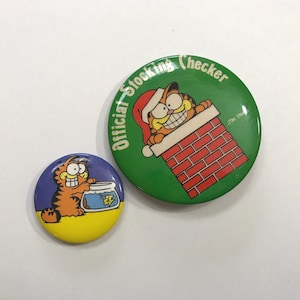 May include: Two round, colourful, pin-back buttons featuring the cartoon character Garfield. The larger button is green with white text that reads "Official Stocking Checker" and features Garfield peeking out of a brick chimney. The smaller button is blue with Garfield holding a fishbowl.
