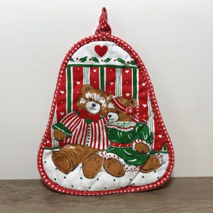 May include: A Christmas-themed oven mitt featuring a red and white polka dot border and a red hanging loop. The mitt showcases a design of two teddy bears in festive attire against a backdrop of red and green stripes and hearts.