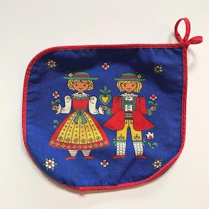 May include: A blue fabric pot holder with a red trim features a folk art design of two figures in traditional clothing. The figures are holding flowers and a heart. The background is filled with white flowers.