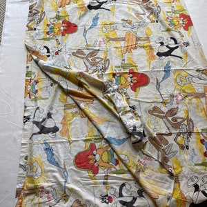 May include: A white cotton fabric with a repeating pattern of cartoon characters from the Looney Tunes show. The characters include Wile E. Coyote, Road Runner, and Sylvester the Cat. The fabric is yellow, orange, and blue with a white background.