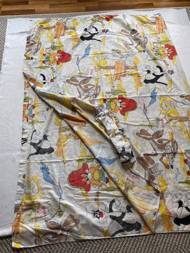 May include: A white cotton fabric with a repeating pattern of cartoon characters from the Looney Tunes show. The characters include Wile E. Coyote, Road Runner, and Sylvester the Cat. The fabric is yellow, orange, and blue with a white background.