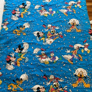 May include: A blue and white cotton fabric with a repeating pattern of Mickey Mouse, Minnie Mouse, Donald Duck, Goofy, and Pluto characters throwing pillows. The characters are all wearing their signature clothing and are surrounded by white confetti.