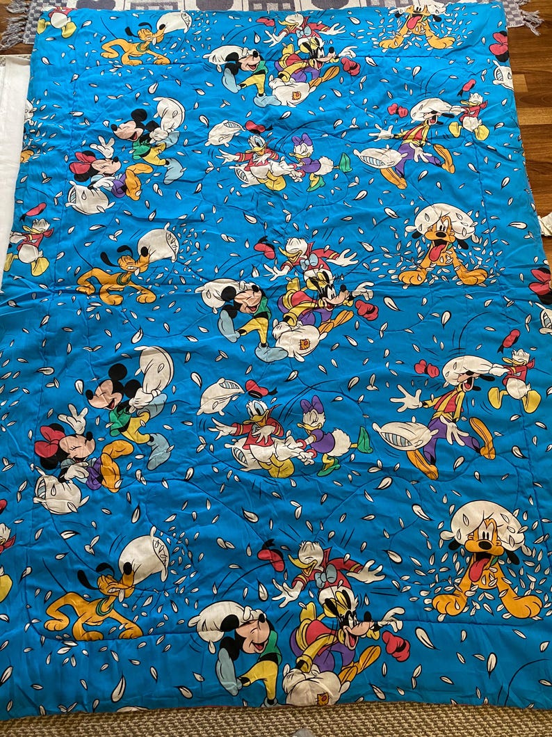 May include: A blue and white cotton fabric with a repeating pattern of Mickey Mouse, Minnie Mouse, Donald Duck, Goofy, and Pluto characters throwing pillows. The characters are all wearing their signature clothing and are surrounded by white confetti.