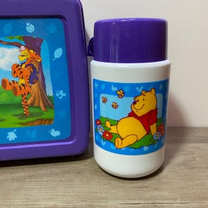 May include: A purple lunchbox with a Winnie the Pooh design and a matching purple and white thermos with a Winnie the Pooh design.
