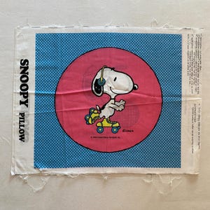 May include: A white fabric panel with a blue polka dot background. A pink circle with a white outline contains a cartoon image of Snoopy wearing headphones and roller skates. The text "SNOOPY PILLOW" is printed vertically on the left side of the panel. The text "SCHROEDER" is printed below Snoopy. The text "1988 United Feature Syndicate, Inc." is printed below the text "SCHROEDER".