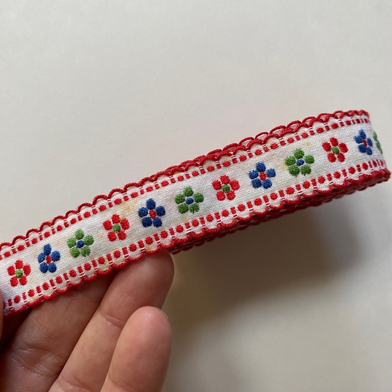 May include: White fabric ribbon with a red scalloped edge and a repeating pattern of red, blue, and green flowers.