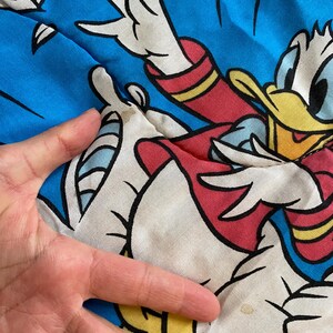 May include: A white and blue fabric with a cartoon image of Donald Duck in a red and yellow outfit. Donald Duck is flying through the air with his arms outstretched. The fabric has a faded appearance and a small yellow stain.