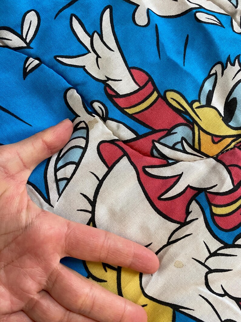 May include: A white and blue fabric with a cartoon image of Donald Duck in a red and yellow outfit. Donald Duck is flying through the air with his arms outstretched. The fabric has a faded appearance and a small yellow stain.