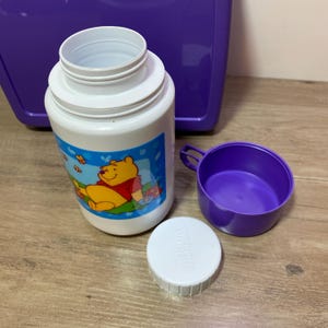 May include: A white Winnie the Pooh thermos with a purple lid and a matching purple cup. The thermos has a picture of Winnie the Pooh sitting on a blue and green background with yellow butterflies.