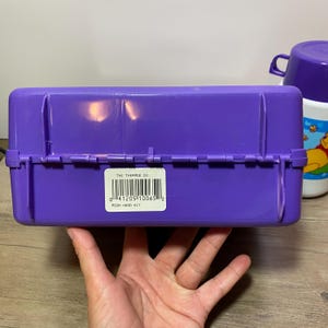 May include: Purple plastic lunchbox with a Winnie the Pooh hard kit label and a barcode. The label reads "The Thermos Co. Pooh Hard Kit".