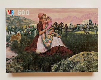 May include: MB 500-piece puzzle featuring a Western-themed illustration. A woman in a bonnet holds a child wrapped in a quilt. The background depicts a wagon train, horses, and a dog near a stream. The puzzle box has a pink, brown, and green color scheme.