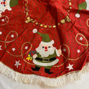 K&ouml;nnte beinhalten: Roter Weihnachtsbaumrock mit einem sich wiederholenden Muster aus winzigen gr&uuml;nen Bl&auml;ttern und goldenen Akzenten. Der Rock hat eine Santa Claus Applikation, wei&szlig;e Sterne und eine wei&szlig;e Fransenborte. Eine festliche Weihnachtsdekoration.