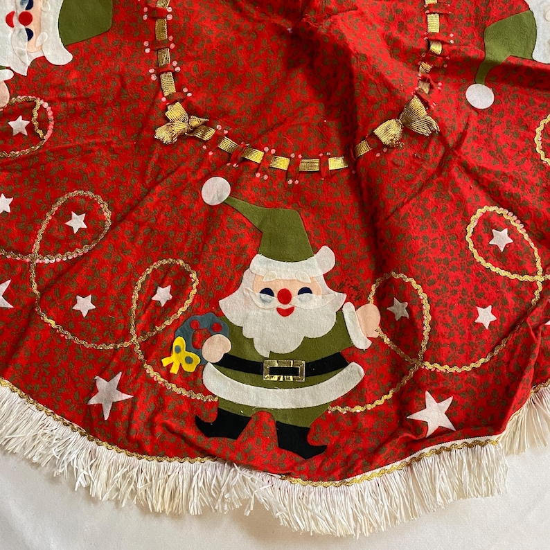 K&ouml;nnte beinhalten: Roter Weihnachtsbaumrock mit einem sich wiederholenden Muster aus winzigen gr&uuml;nen Bl&auml;ttern und goldenen Akzenten. Der Rock hat eine Santa Claus Applikation, wei&szlig;e Sterne und eine wei&szlig;e Fransenborte. Eine festliche Weihnachtsdekoration.