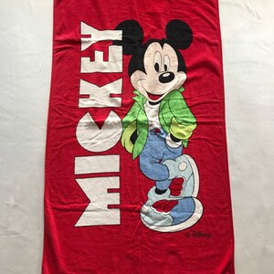 May include: Red beach towel featuring Mickey Mouse in a green jacket, blue jeans, and white shoes. The word "MICKEY" is printed vertically in white. The Disney logo is in the bottom right corner.