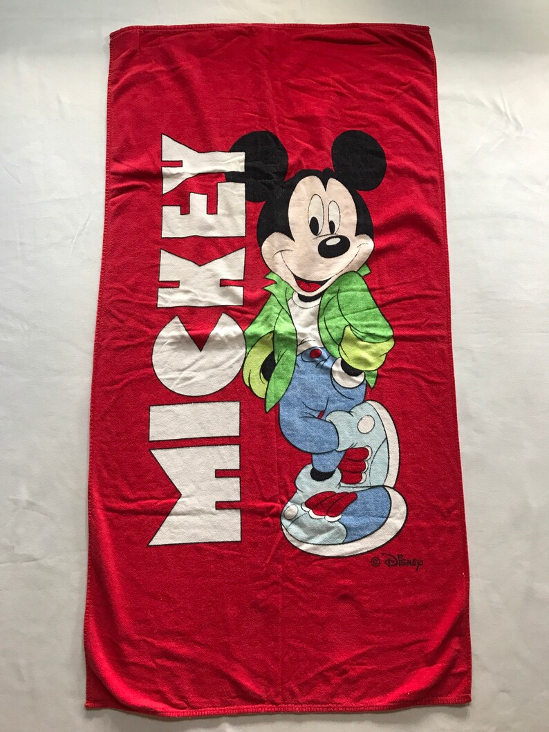 May include: Red beach towel featuring Mickey Mouse in a green jacket, blue jeans, and white shoes. The word "MICKEY" is printed vertically in white. The Disney logo is in the bottom right corner.