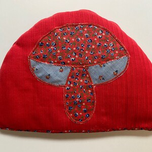 May include: Red fabric tea cosy shaped like a mushroom with a floral pattern. The mushroom is made of patchwork fabric with blue and red floral patterns.