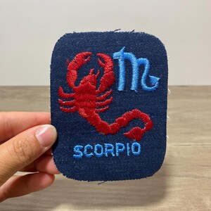 以下が含まれることがあります： 紺色の長方形の刺繍ワッペン。赤いサソリのグラフィック、水色の蠍座のシンボル、そして水色の大文字で「SCORPIO」という文字が刺繍されています。ワッペンは角が丸く、布地のような質感の表面をしています。