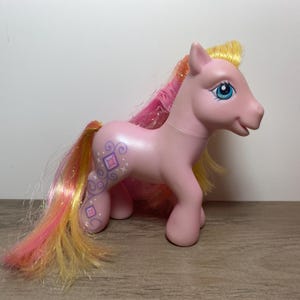 May include: Pink My Little Pony toy with a yellow, pink, and orange mane and tail. The pony has a blue eye and a swirl design on its side with pink and purple diamonds.