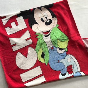 May include: Red Mickey Mouse towel featuring a smiling Mickey in a green jacket, blue jeans, and white shoes. The word "MICKEY" is printed in large white letters on the towel. A fun and colorful item for Disney fans.