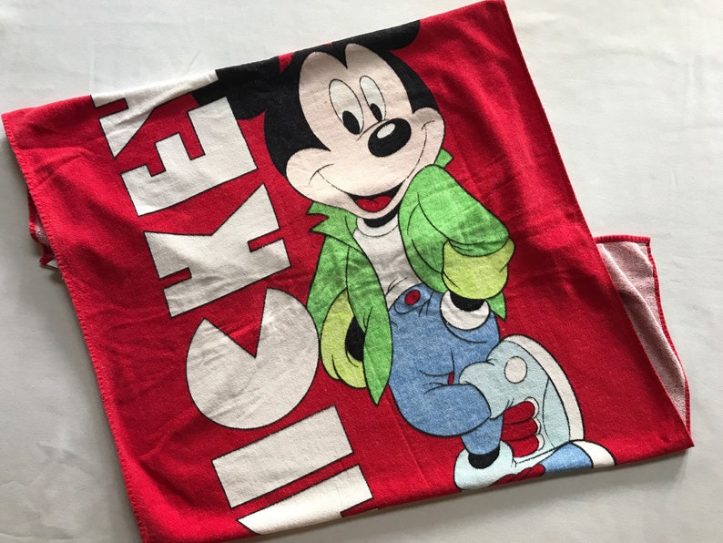 May include: Red Mickey Mouse towel featuring a smiling Mickey in a green jacket, blue jeans, and white shoes. The word "MICKEY" is printed in large white letters on the towel. A fun and colorful item for Disney fans.