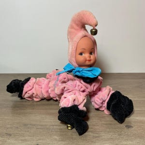 Peut inclure: Une poupée vintage avec un corps en velours rose et un chapeau assorti avec une petite cloche. La poupée a un visage en plastique, un nœud bleu et des pieds en velours côtelé noir. La poupée est sur une surface en bois.