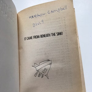 Op de afbeelding: Een boek getiteld "It Came From Beneath The Sink!" met een handgeschreven inscriptie die "Matthew Campbell Div. 9" luidt. Op de pagina is ook een eenvoudige tekening van een schepsel te zien.