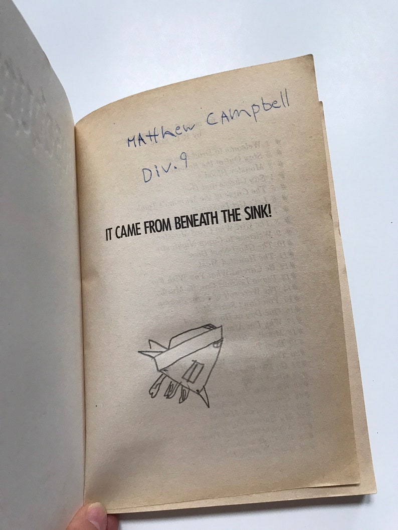 Op de afbeelding: Een boek getiteld "It Came From Beneath The Sink!" met een handgeschreven inscriptie die "Matthew Campbell Div. 9" luidt. Op de pagina is ook een eenvoudige tekening van een schepsel te zien.