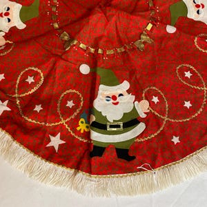 K&ouml;nnte beinhalten: Roter Weihnachtsbaumrock mit einem sich wiederholenden Muster aus kleinen gr&uuml;nen Bl&auml;ttern. Der Rock hat eine Santa Claus Applikation, wei&szlig;e Sterne und goldene Verzierungen. Der Rand ist mit einer wei&szlig;en Fransen versehen.