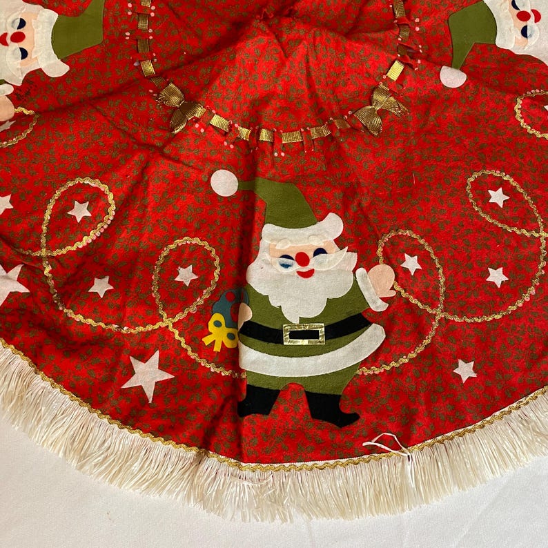 K&ouml;nnte beinhalten: Roter Weihnachtsbaumrock mit einem sich wiederholenden Muster aus kleinen gr&uuml;nen Bl&auml;ttern. Der Rock hat eine Santa Claus Applikation, wei&szlig;e Sterne und goldene Verzierungen. Der Rand ist mit einer wei&szlig;en Fransen versehen.