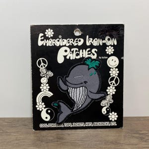 May include: A gray whale embroidered iron-on patch on a black backing card with white text reading Embroidered Iron-On Patches by kalan and Cool for jeans, tops, jackets, hats, backpacks, etc. The card features white flower, peace sign, and butterfly icons.