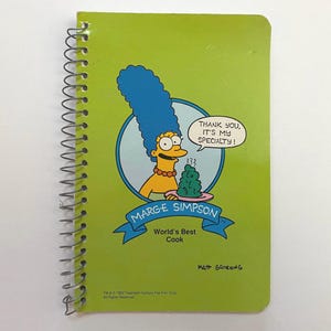 May include: A green spiral notebook with a cartoon image of Marge Simpson holding a plate of food. The text on the notebook says "Marge Simpson, World's Best Cook" and "Thank you, it's my specialty!"