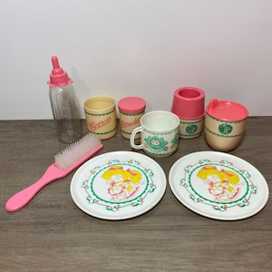May include: A collection of plastic toy nursery accessories including a clear bottle with a pink cap, a pink hairbrush, two white plates with a yellow-haired doll graphic, a mug, and containers labeled Cotton and Powder with pink lids.